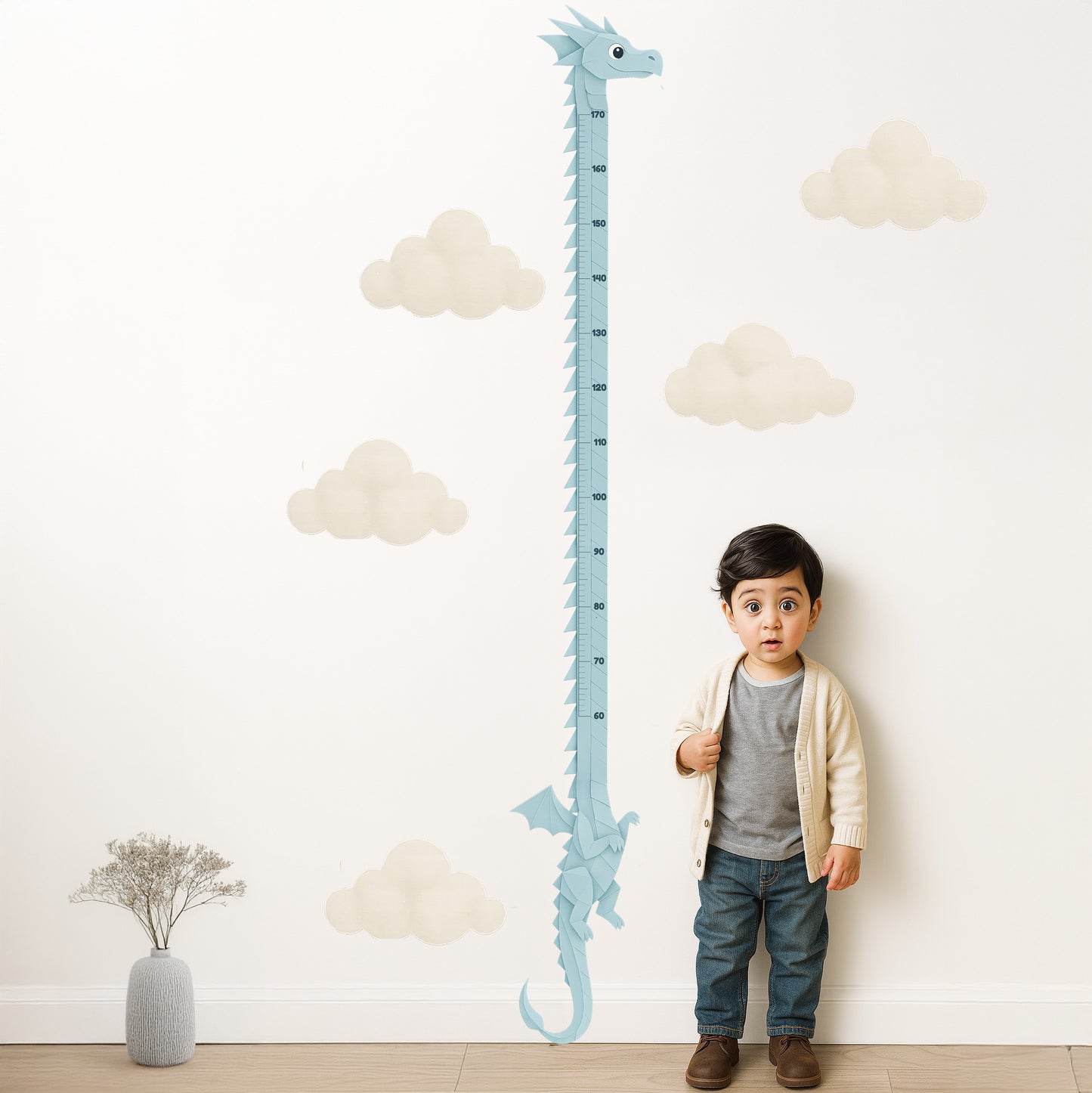 A young South Asian boy stands indoors beside a tall, pastel blue dragon-themed height chart on a cream wall with soft paper-cut clouds. The dragon's long, folded-paper body is marked with height measurements, and the child looks upward with a surprised expression. The floor is wooden, and a small vase with dried flowers sits to the left.